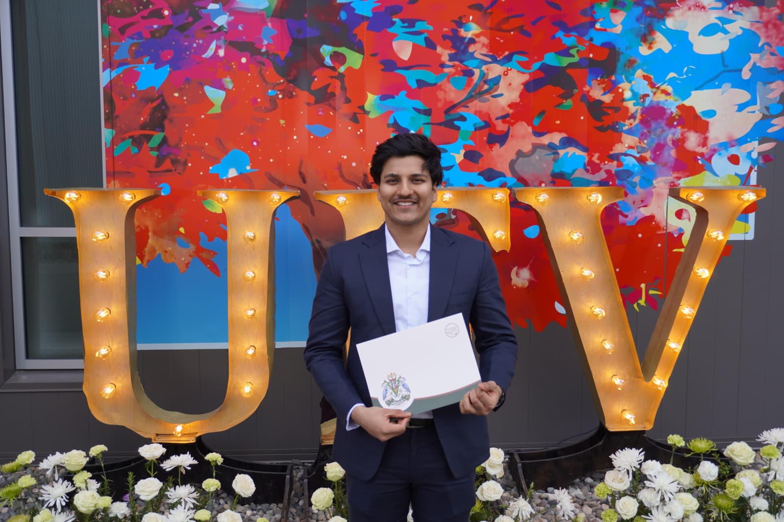 Nishant holding his degree in front of the lit-up UFV letters at convocation